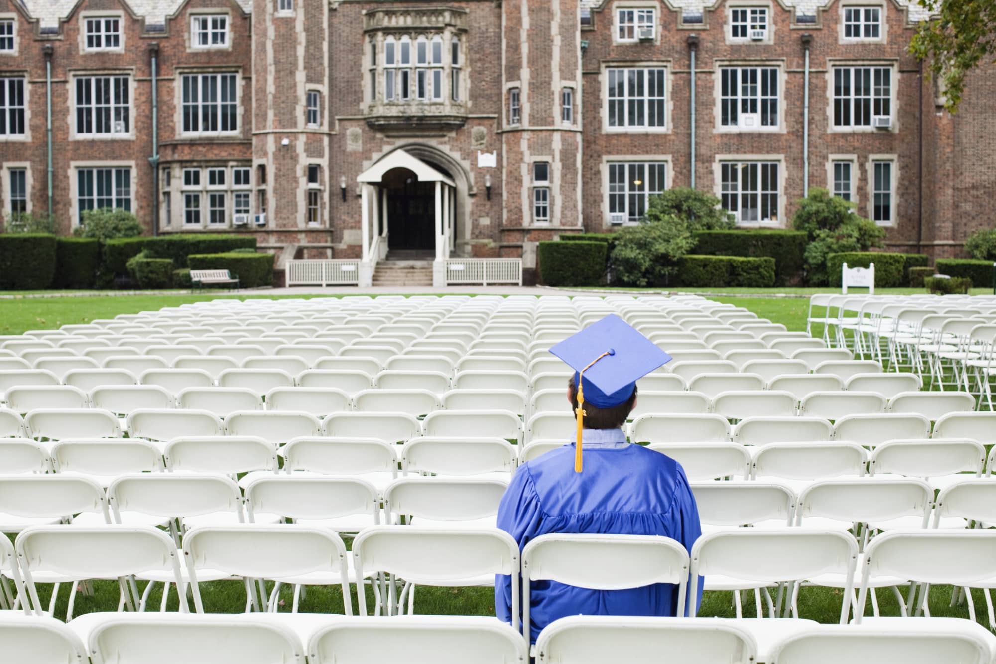 College graduate student wearing a blue cap and gown sits alone in an empty outdoor auditorium before his graduation ceremony.