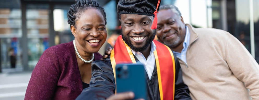 Excited Black male college graduate taking selfie photos with his proud parents after his graduation ceremony. He is wearing a black cap and gown and holding his degree in his left hand. He is using his right hand to take photos with his cell phone.