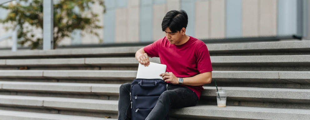 Male Asian American college student sits on stairs outside of a university campus building during his break. He is packing away his laptop in his backpack before heading off to his next class.