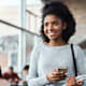Black female college student smiles while standing in a student center on campus. She is holding notebooks in her left hand while using her right hand to check emails on her mobile phone.