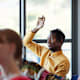 Black male community college student sitting at a desk in a classroom with other students. His arm is raised to answer a question from the professor off-screen.