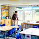 Caucasian male elementary school teacher walking around student desks in a classroom. He is handing out class workbooks before students arrive in the morning.