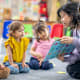 A preschool teacher sits on the floor of her classroom with a small group of students as she reads them a book.
